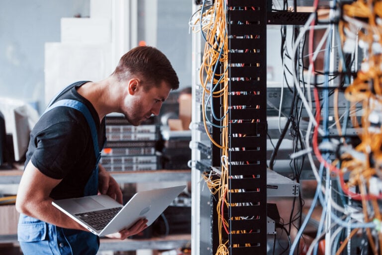 Young man in uniform and with laptop works with internet equipment and wires in server room